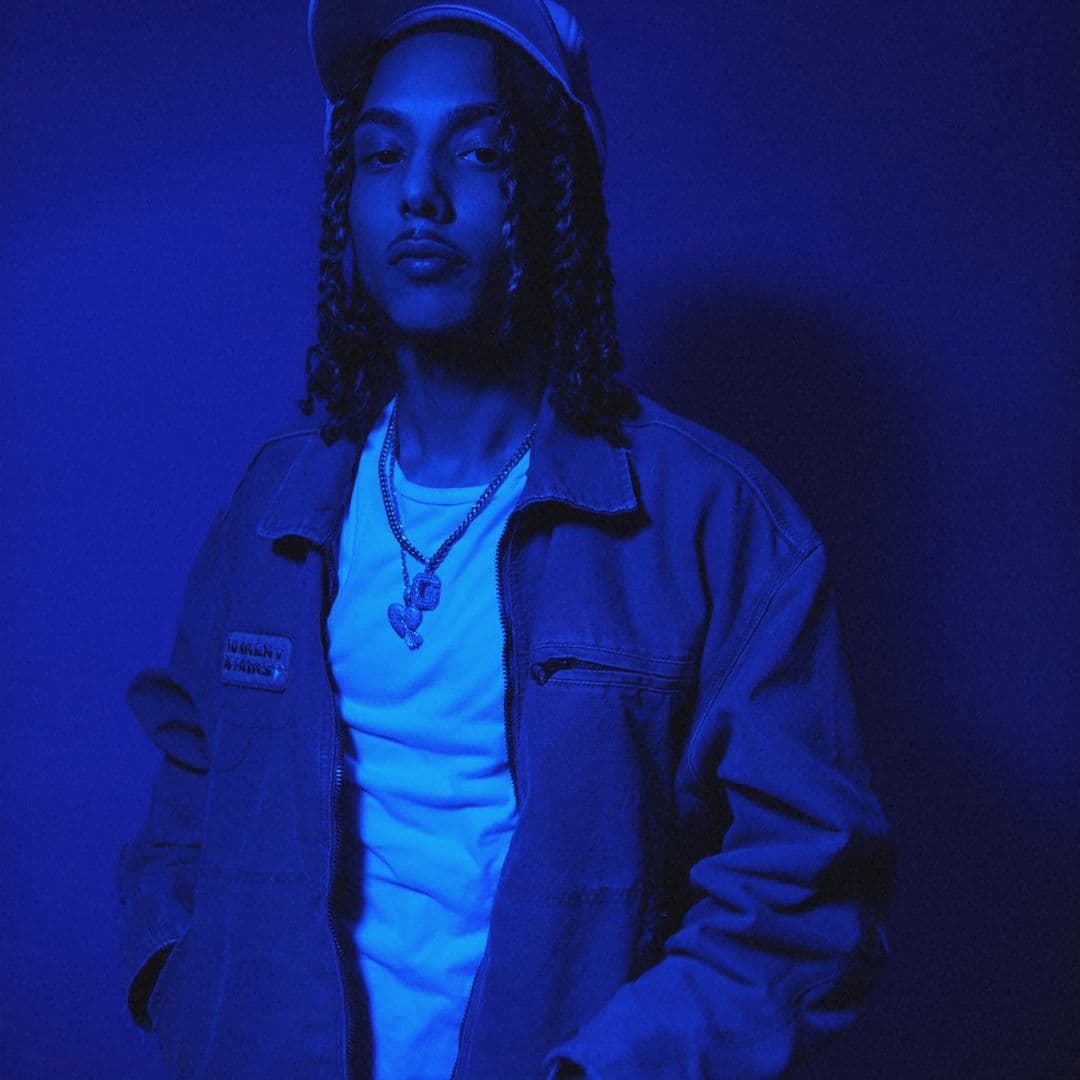 Jayggm in a denim jacket and chain, bathed in deep blue light