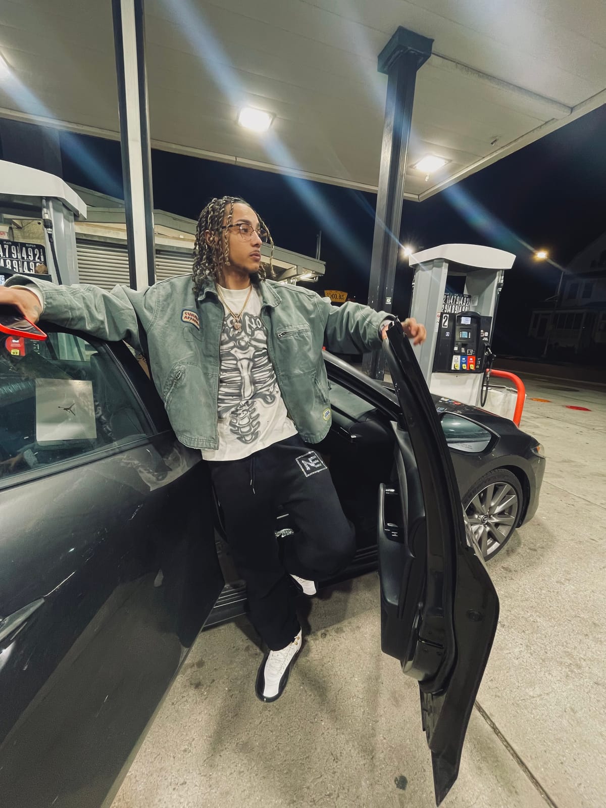 Jayggm leaning on a car door at a gas station at night